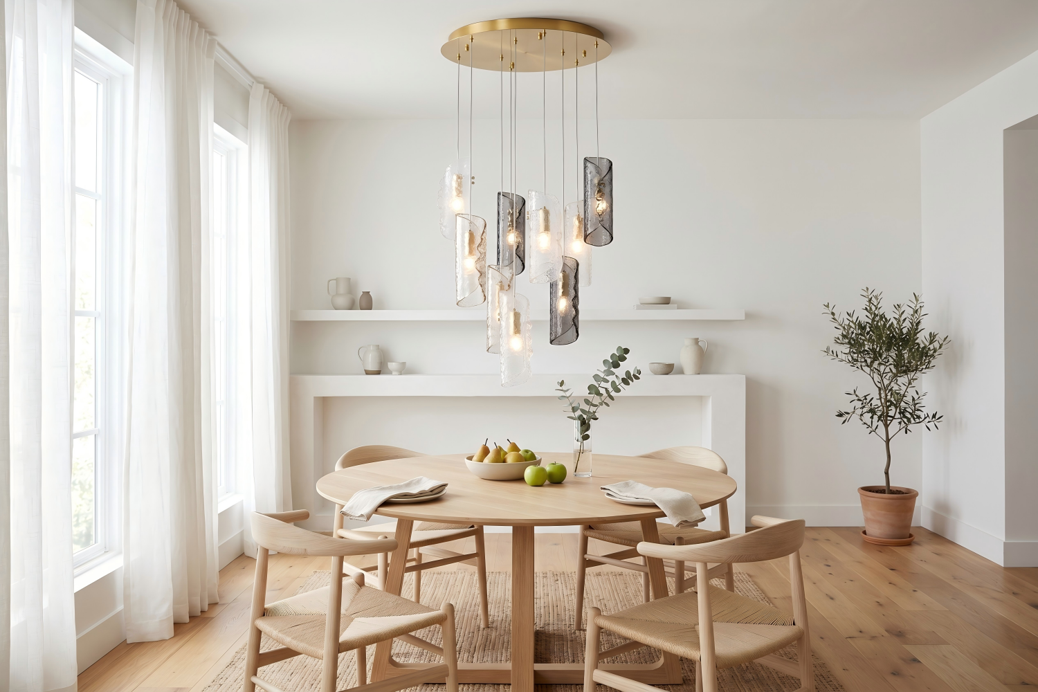 Cascading textured glass chandelier with 9 staggered tube shades in clear and smoky grey glass and polished brass canopy, hanging above a round white oak dining table in a bright Scandinavian-style dining room