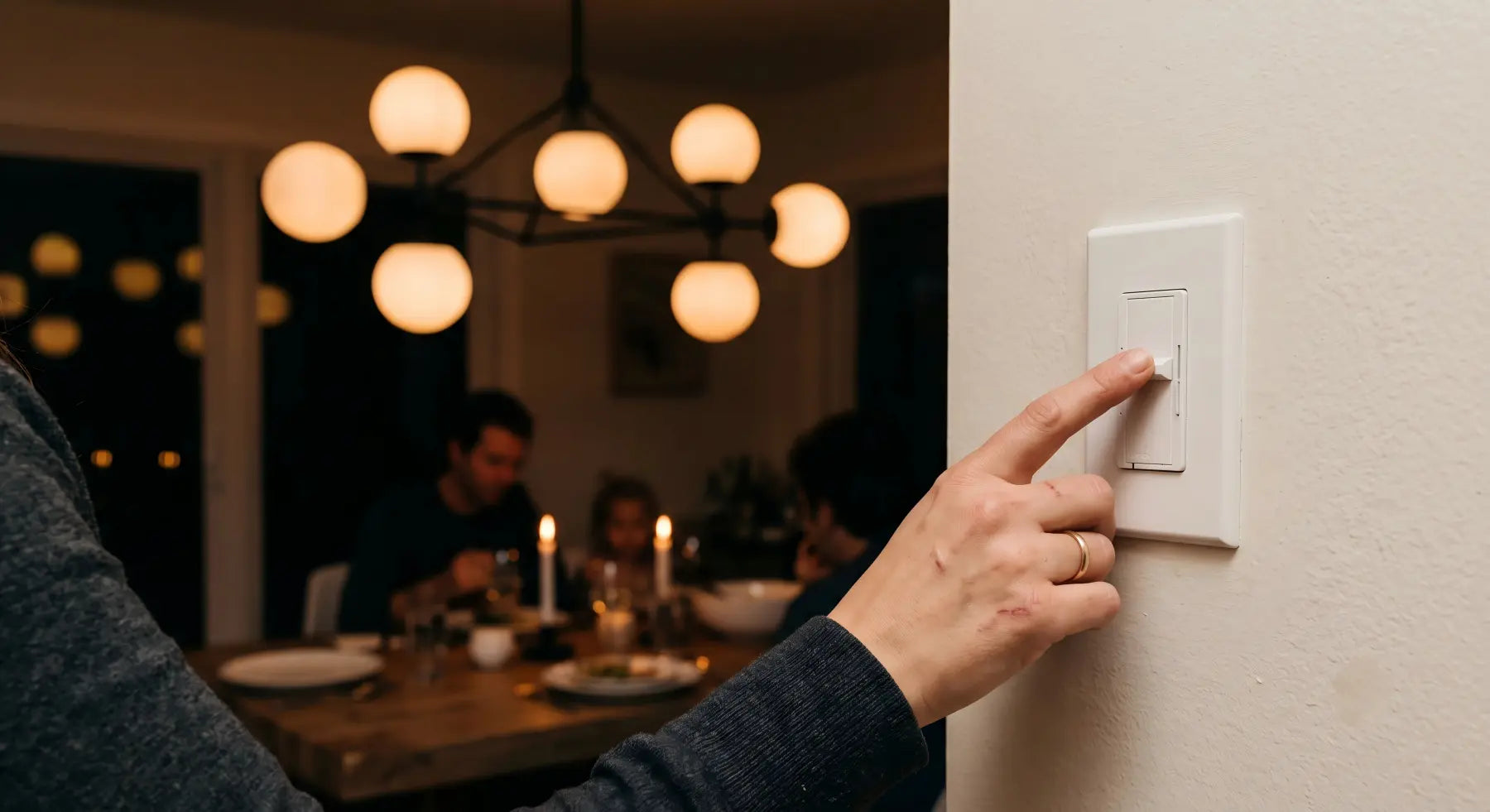 Hand adjusting a wall dimmer switch with a warmly dimmed chandelier glowing in the background